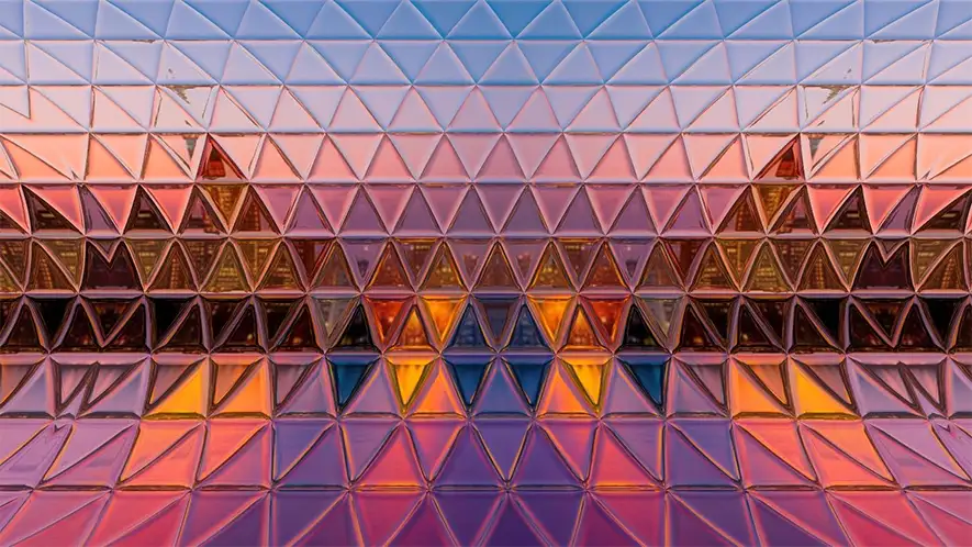 Mosaikartige Glasfassade mit dreieckigen Scheiben in verschiedenen Farben bei Sonnenuntergang.