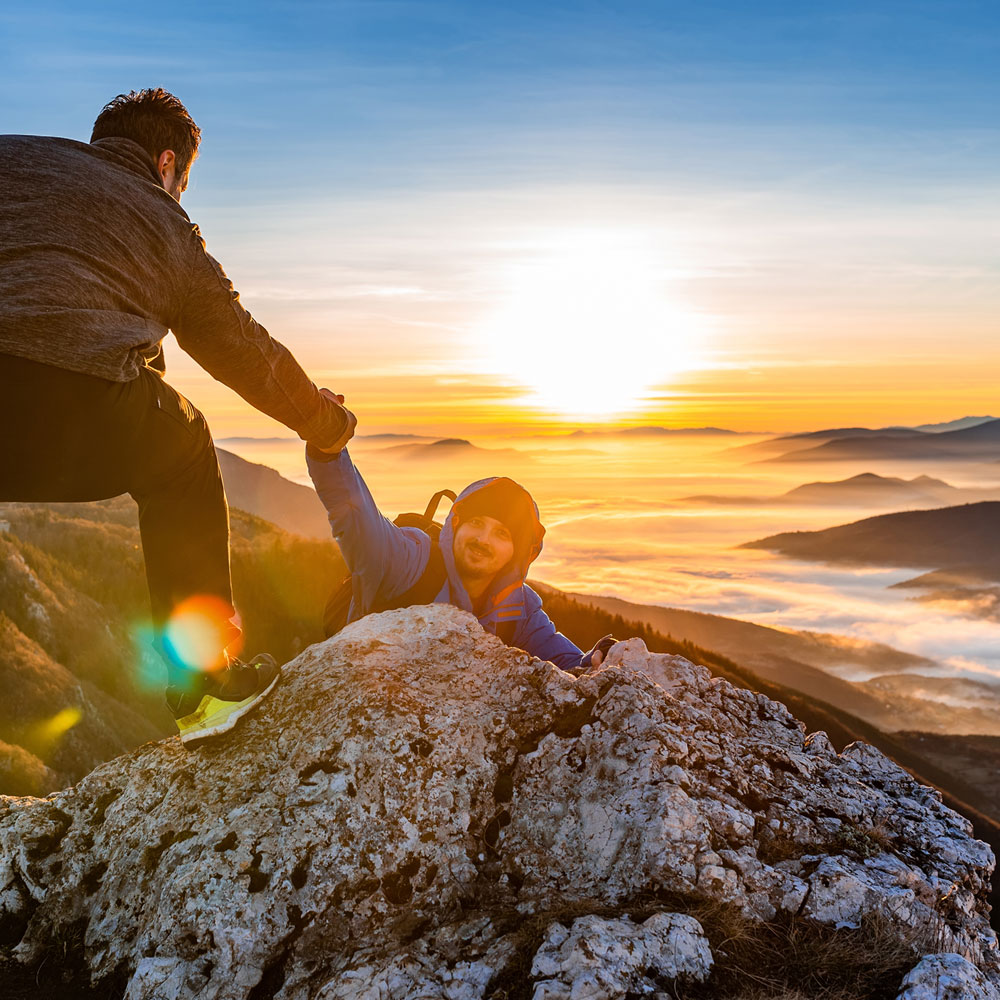 Zwei Wanderer an einem Berggipfel beim Sonnenaufgang, einer hilft dem anderen beim Hochziehen über einen Felsen.