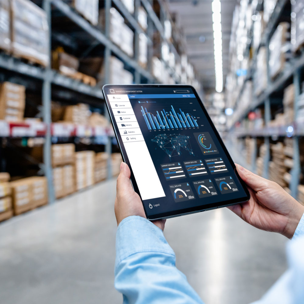 Eine Person hält ein Tablet in den Händen, auf dessen Bildschirm eine Lagerverwaltungssoftware mit Diagrammen, Karten und Datenanzeigen sichtbar ist. Im Hintergrund ist ein weitläufiges Lager mit vielen Regalen und Kartons zu sehen.