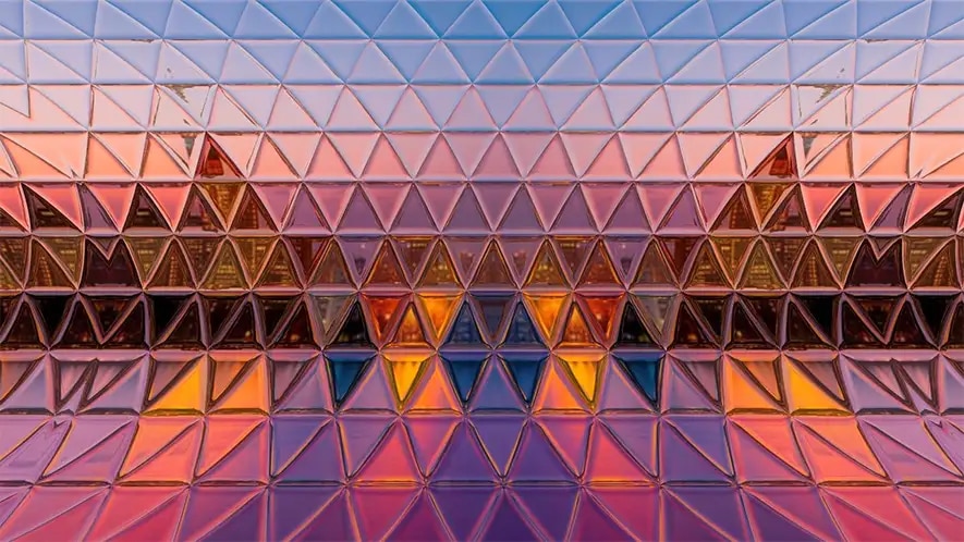 Glass wall with triangular, reflective segments mirroring a colorful, abstract pattern of pink, purple, orange, and blue hues at sunset