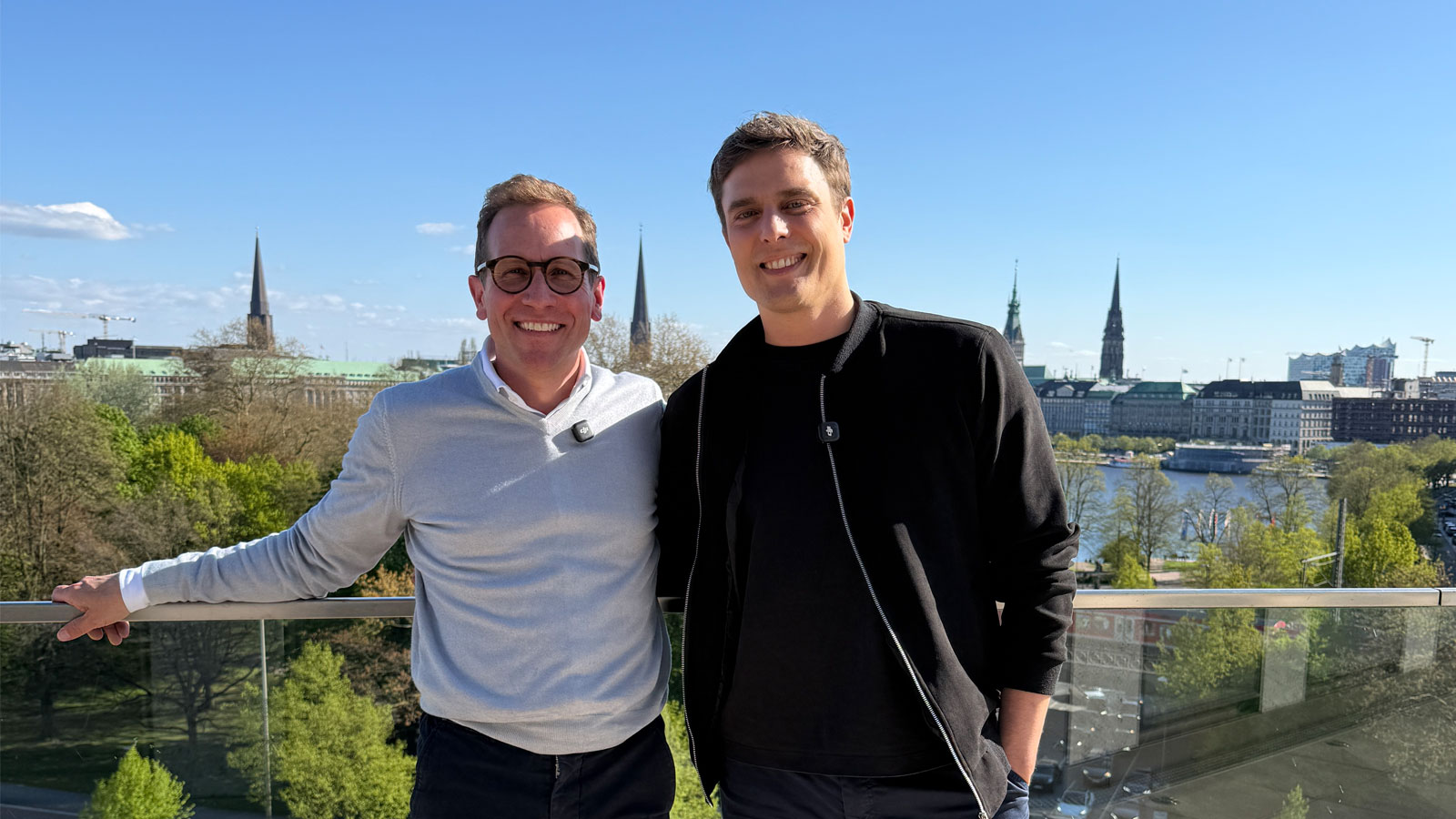 Christian Kaspar and Constantin Schreiber standing side by side on a glass balcony terrace overlooking a body of water and a city with several church spires; faces obscured.