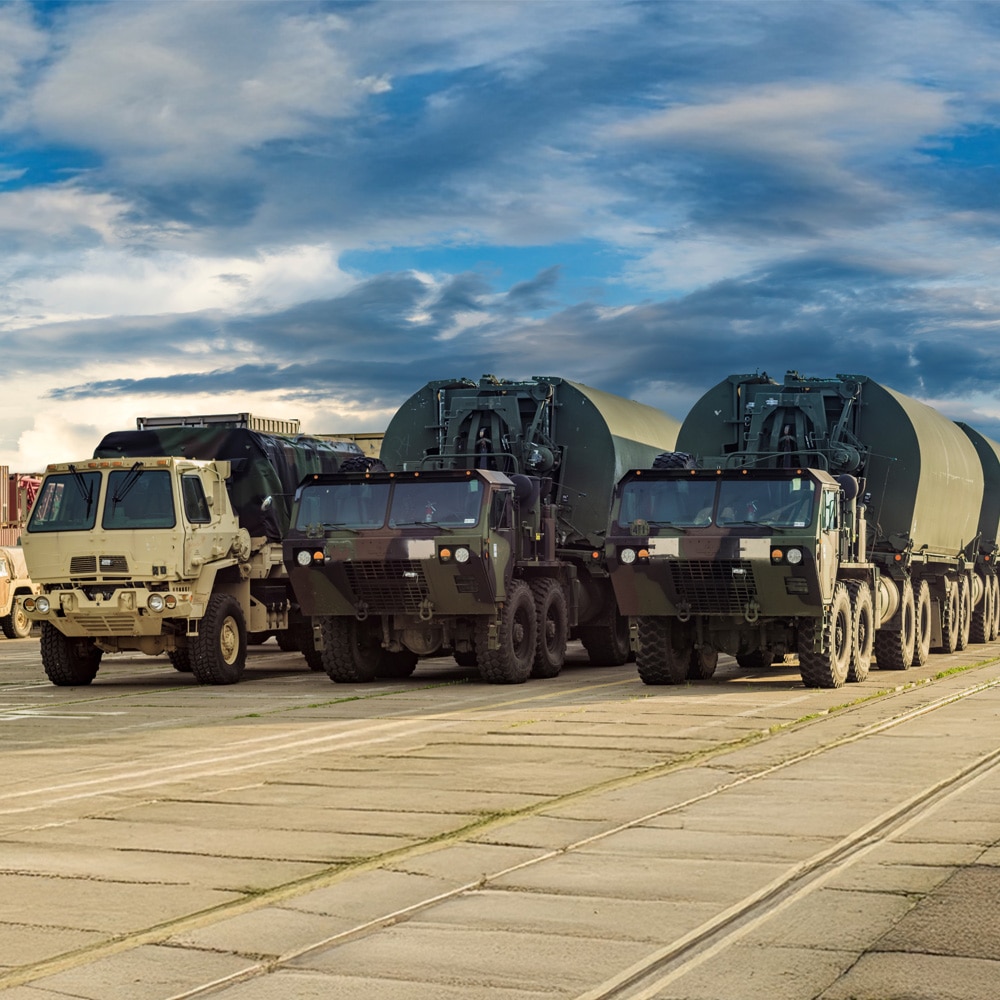 Several military vehicles with large cylindrical trailers on a concrete lot under partly cloudy skies.