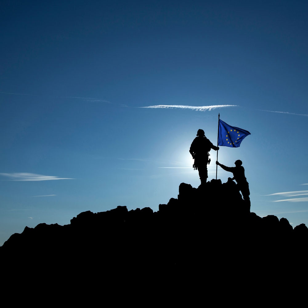 Silhouettes of two soldiers on a mountaintop with a waving European Union flag.