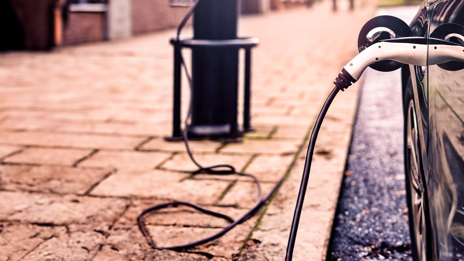 A black electric car is parked and being charged via a cable connected to a modern public charging station on a paved street.