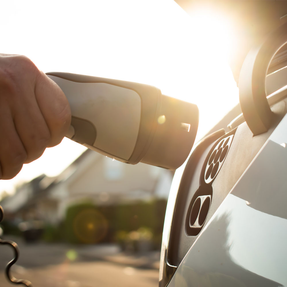 Hand holding a charging connector and plugging it into an electric car's charging port, with a sunny street in the background.
