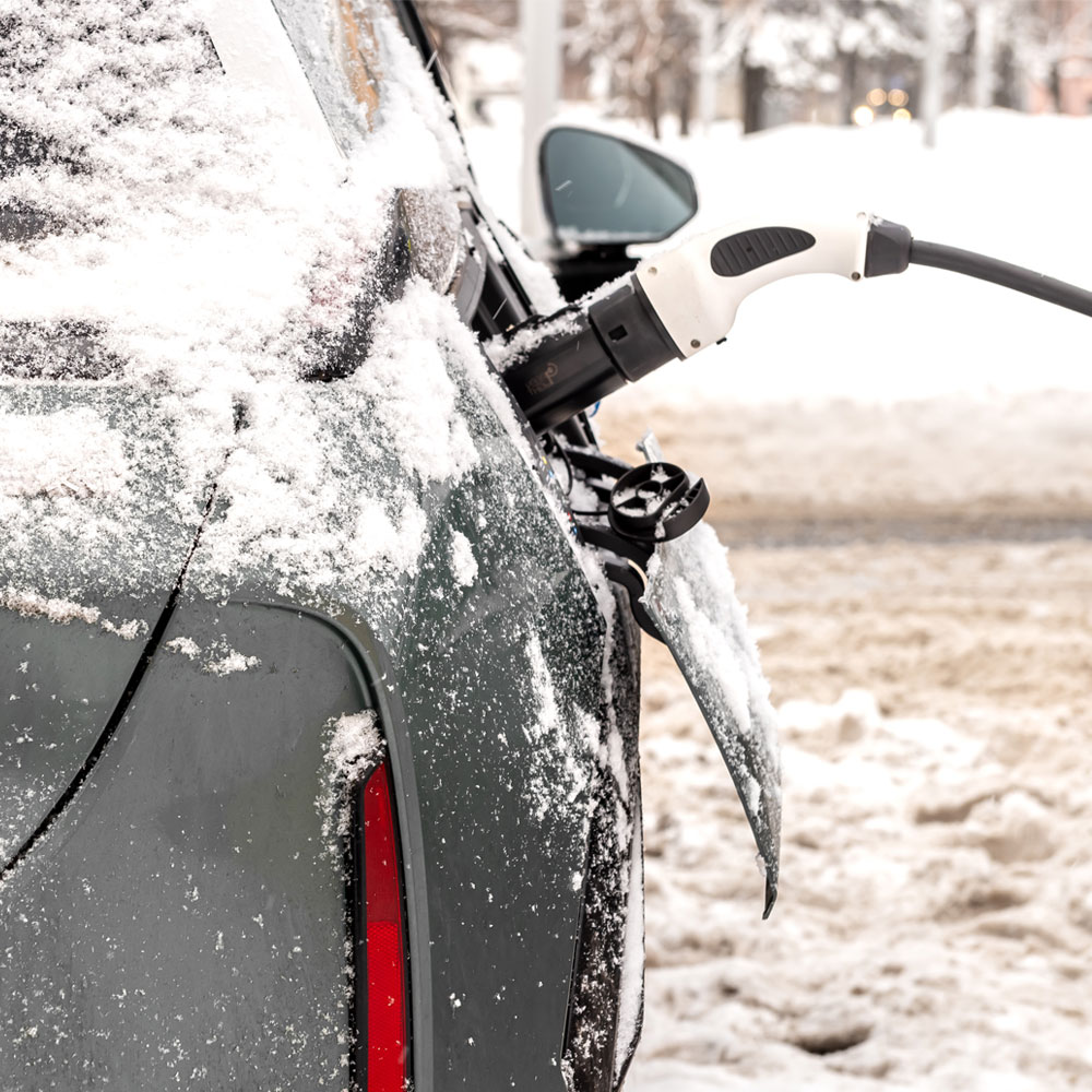 Electric car being charged at a charging station in snowy conditions, showing the snow-covered rear and a charging plug inserted into the charging port.