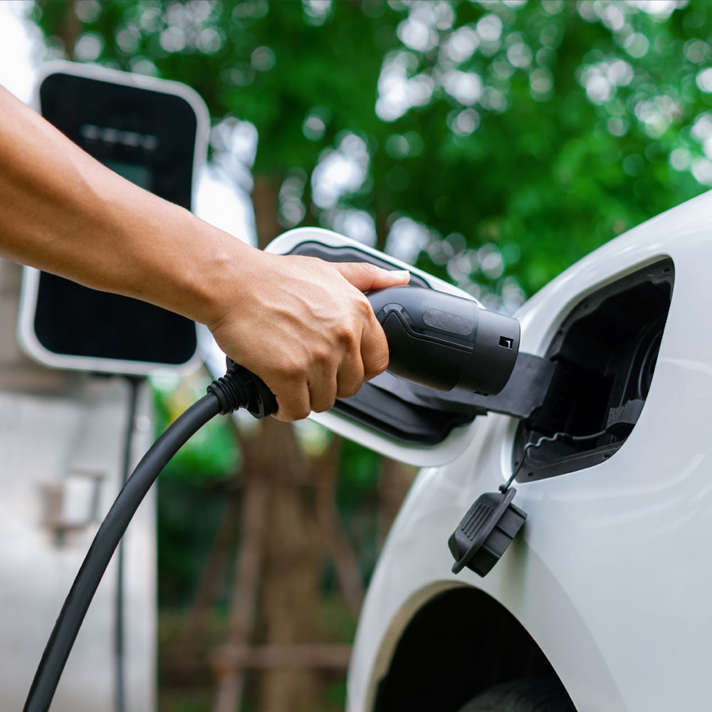 Hand holding a charging plug and inserting it into a white electric car at a charging station.