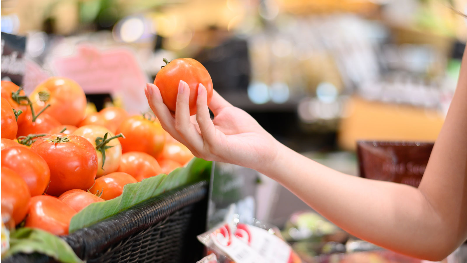 A hand is holding a fresh, red tomato above a basket filled with more tomatoes in a supermarket.