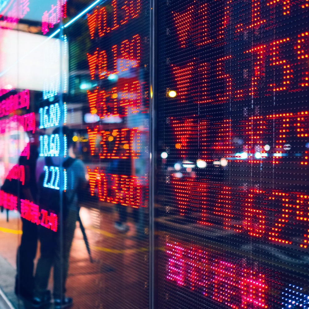 Electronic stock market board with red and blue numbers and percentages, reflected on a glass facade, with blurred pedestrians in the background.