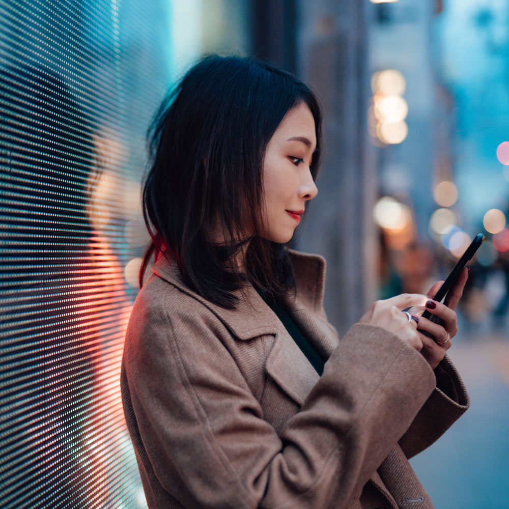 Person with pixelated face leans against a glowing LED wall and taps on a smartphone; blurred city lights in the background.