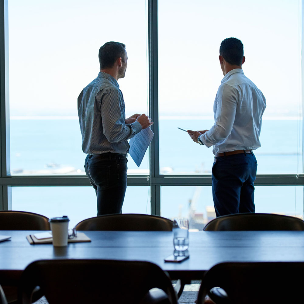 Two people in business attire stand with their backs to the camera at large panoramic windows, looking out over a harbor; a conference table with a laptop, coffee cup and glass is in the foreground.