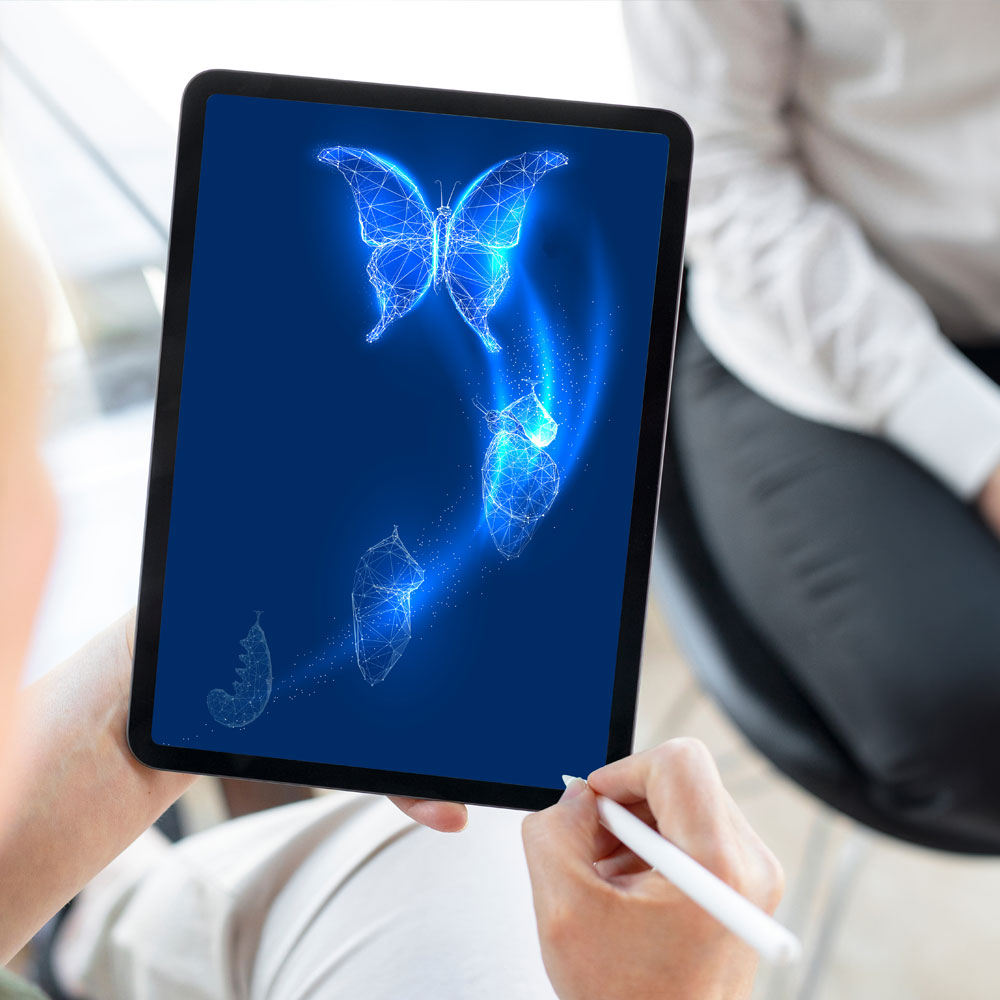 Tablet held portrait shows a glowing digital sequence from caterpillar to chrysalis to butterfly on a deep blue screen; a person holds the tablet and touches the display with a white stylus, another person sits blurred in the background.