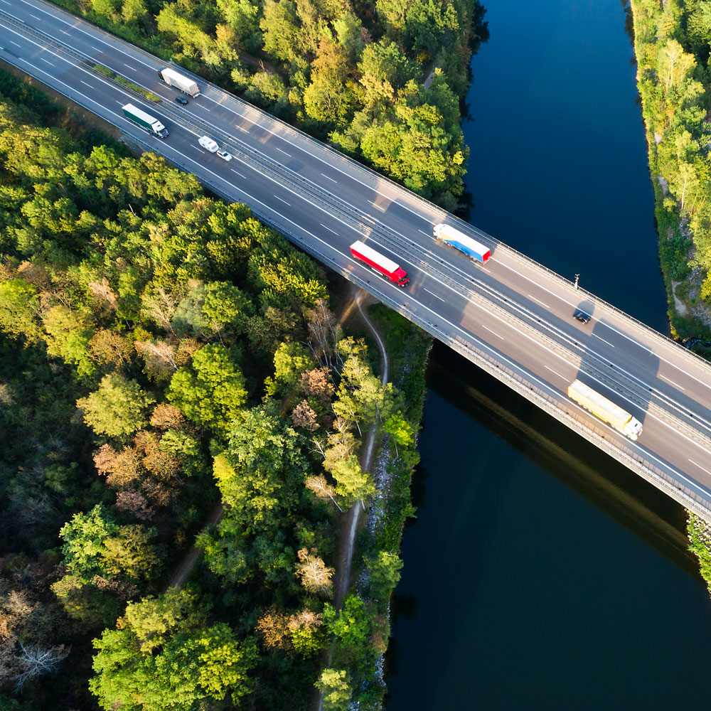 Aerial view of a multi-lane highway bridge crossing a river, surrounded by dense forest, with several cars and trucks.