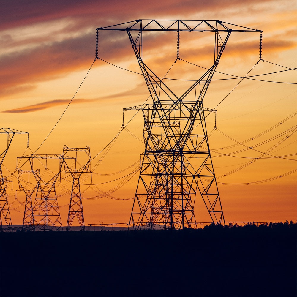 The image shows multiple high-voltage pylons stretching in a long row across a landscape. The background sky is colored in warm orange tones and darker blue hues of a sunset.